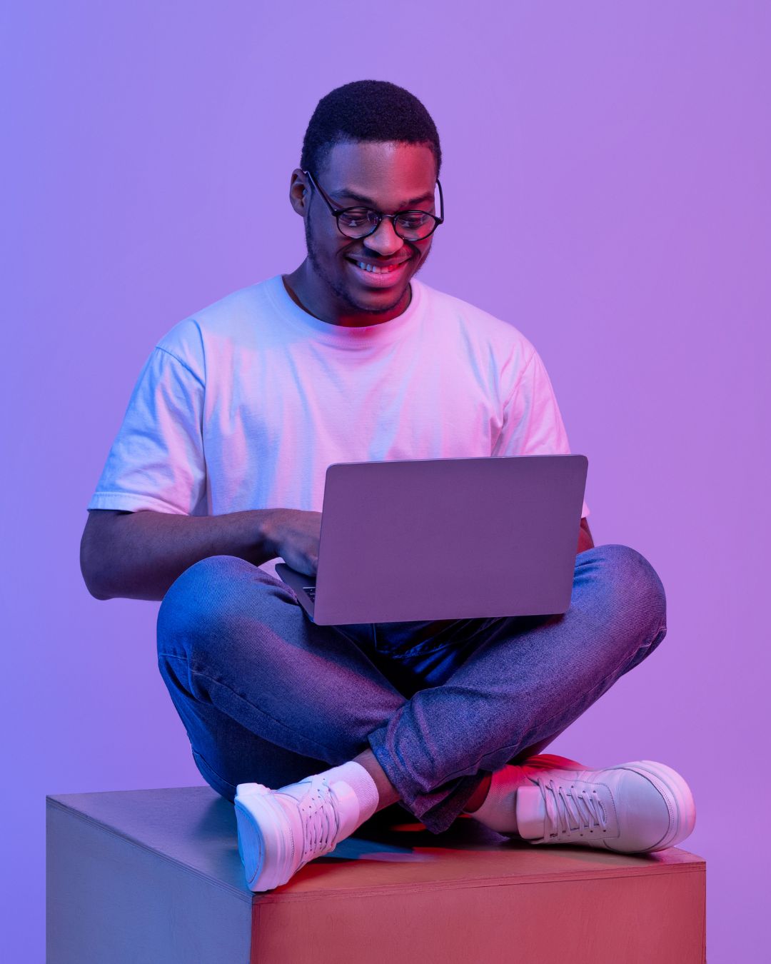 Young Nigerian entrepreneur sitting cross-legged with a laptop on a purple studio backdrop, smiling as sales roll in on BazaarFlow